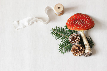 Christmas festive styled stock composition. Red fly agaric mushroom, silk ribbon, pine cones and fir tree branches on white linen background. Winter table flat lay, top view. Decorative still life.