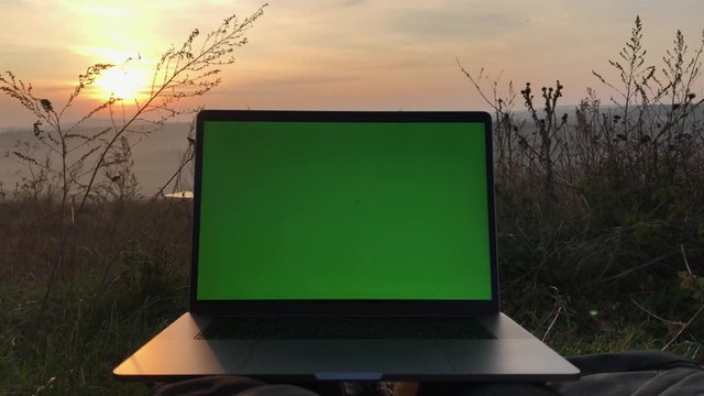 Close-up of modern laptop with green screen on background of sunset.