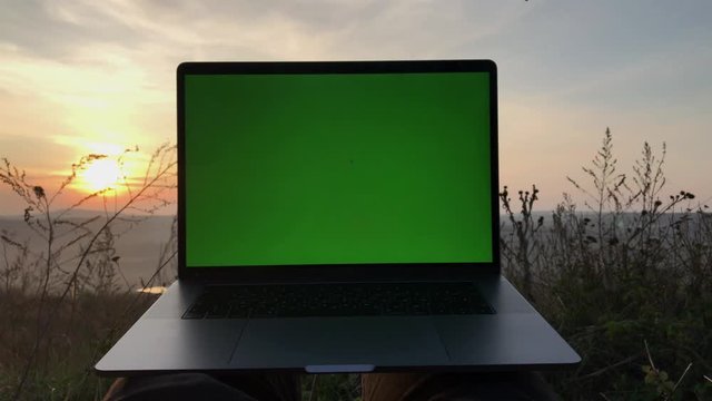 Close-up of modern laptop with green screen on background of sunset.
