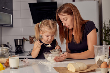 Mother and daughter preparing a sweet cake using flour, milk, sitting on chairs at a table in a modern kitchen. Girl holding a whisk, stirring eggs in a bowl, preparing pancake dough with her mom.