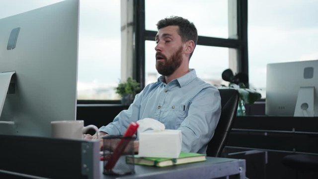 Exhausted allergic young man office worker suffering flu blowing nose with napkins continue working by personal computer. Hardworking people.