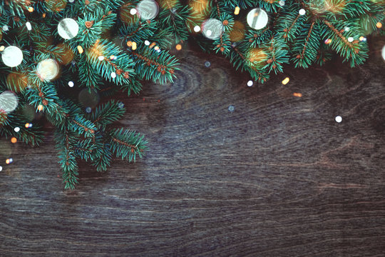 Natural Christmas background with border of fluffy green fir branches on a textured dark brown wood background with christmas lights. Copy space for text, flat lay, top view.