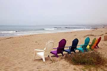 Colored Adirondack chairs face the Pacific Ocean on a sandy beach.