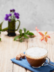 Glass cup of cappuccino on a blue napkin with a spoon and slices of cane sugar. Wood background. Autumn leaves and wild flower in a vase in the background. Close-up.