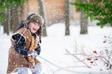 Fototapeta na wymiar Feast of the Maslenitsa. A child with bagels pulls a rope on a winter background. A guy in a cap with earflaps having fun on a winter Russian holiday