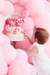 cute little baby girl poking her finger in birthday cake, decorated with pastel pink balloons on...