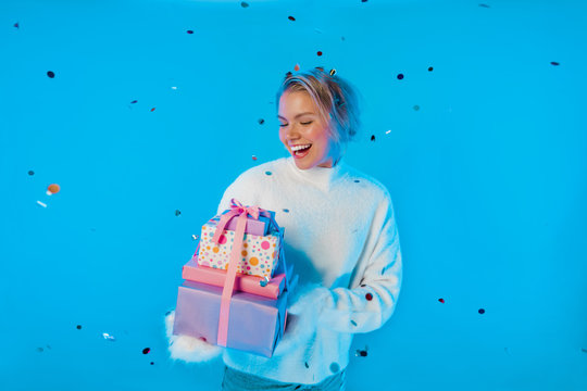 Girl Holding A Gift And Laughing Merrily. Metallic Shiny Confetti Falls From Above. A Blonde Girl In Fluffy Mittens And A White Sweater On A Bright Blue Isolated Neon Background. Neon Party Light. Gif