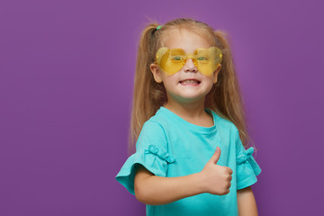 Portrait of little child girl heart shaped sunglasses isolated on purple background. Shows thumbs up, everything is ok. Happy childhood, wow and shock concept
