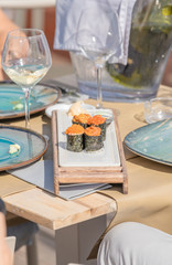 Naklejka na ściany i meble Delicious sushi served on a kind of food chopping board placed on a table with glasses of vin behind and the background out of focus.