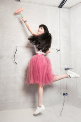 Young woman with pink chiffon skirt and sneakers jumping in front of a gray wall with paint roller...