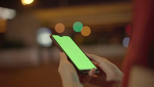 Young Traveler Asian Woman Using Application For Calling Taxi Getting To Home By Mobile Phone With Green Mock Up Screen On Street Night In City. Student Girl Chatting Online While Relax. Chroma Key.