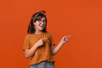 Fototapeta premium Smiling brunette woman in a t-shirt, headband and eyeglasses pointing with her fingers at the right side isolated over orange background.