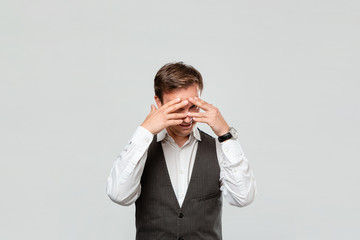 Handsome man in a white shirt and grey vest hiding behind his hands isolated over grey background.