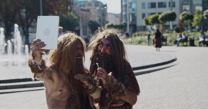 Neanderthals in animal fur using modern tablet take selfie browsing internet fun in modern city center caveman civilization computer evolution future homo sapiens human hunter-gatherers slow motion