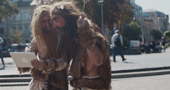 Wonder neanderthals in animal fur using modern tablet browsing internet having fun in city center caveman civilization computer device evolution future homo sapiens human hunter-gatherers slow motion