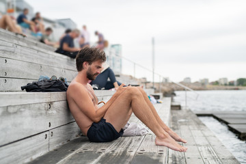 Man sitting on wooden steps and checking smart phone after swimming in the ocean