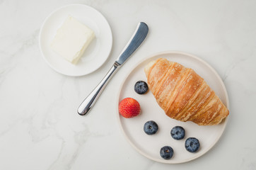 Croissant with berries in white bowl and butter knife on white stone background. Top view