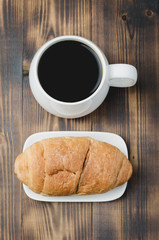Naklejka premium White mug with black coffee and croissant on wooden background. Top view. Coffee break.