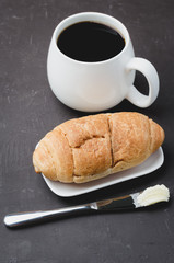 French breakfast. White mug with black coffee and croissant with butter knife on dark stone table. Coffee break.