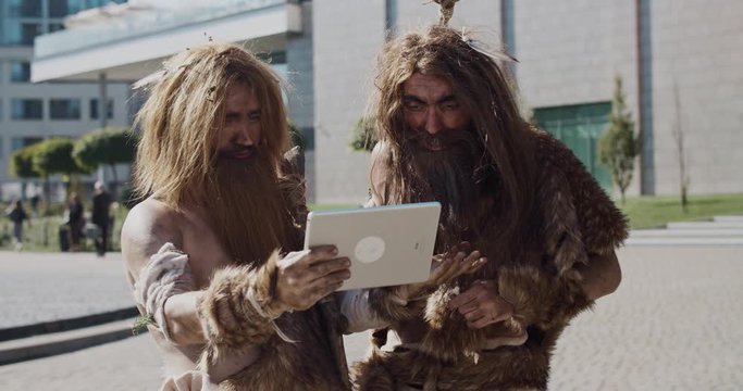 Caveman in animal fur using modern tablet browsing internet having fun civilization computer device evolution future homo sapiens humanity hunter-gatherers neanderthals paleolithic slow motion