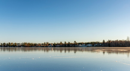Obraz premium The swedish village lays far away, beside the big crystal clear frozen lake. The surface looks like huge mirror, clear skies, very cold winter day, Northern Sweden
