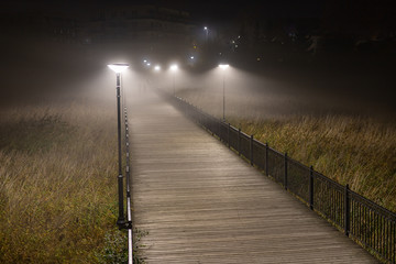 Wooden footbridge illuminated by the light of a lantern in the fog. Passage over wetlands at night in the fog.