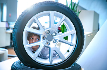 Little boy looking through a tire with a car in a car salon. Salesman in modern car showroom.