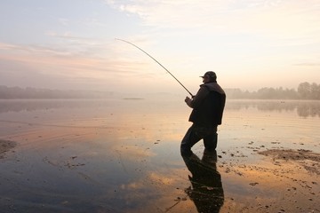 fisherman standing in a lake and catching the fish during foggy dawn