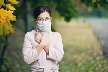 a woman in a medical mask, coat and glasses presses her hands to her chest.