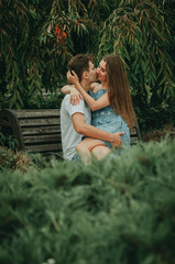 Fototapeta premium Beautiful couple in love sits in a park on a bench in the summer, hugs and kisses. The girl sits on the guy’s lap.