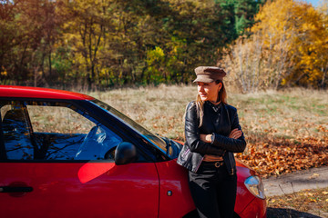 Young woman standing by car on autumn road. Driver stopped auto in forest to enjoy fall landscape