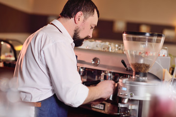 a handsome barista man in a white shirt and apron prepares fragrant coffee on the background of a coffee shop and a coffee machine