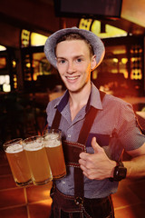 a young attractive man in traditional Bavarian clothes and a hat with a feather holds in his hands a lot of glasses of beer on the pub background during the celebration of the Oktoberfest