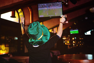 a young man with a mug of dark beer and a green hat from Oktoberfest watching football in a sports bar or pub.