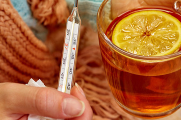 A thermometer is held by a woman's hand next to a Cup of hot tea with a lemon on the background of...