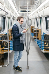 Pretty man with glasses rides alone in the subway train. He is wearing a blue coat, gray scarf and black sneakers. In his hands he holds mobile phone. Digital dependence, social networks addiction.