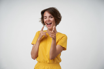 Cheerful young woman with short brown hair wearing yellow t-shirt while standing over white background. winking happily at camera and pointing with raised forefingers