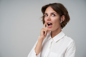 Surprised young beautiful female with short brown hair wearing earphones and looking aside with wide mouth opened, standing over white background in formal clothes
