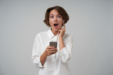 Studio photo of amazed brunette woman with short haircut wearing eyewear and hearing unexpected news, holding smartphone in her hands while posing over white background