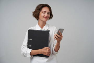 Portrait of pretty brunette woman with short haircut holding folder with documents and typing message on her mobile phone, being in nice mood, isolated over white background