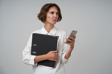Thoughtful young brunette woman with casual hairstyle keeping mobile phone in hand and looking aside thoughtfully, making appointment on working day, posing over white office wall