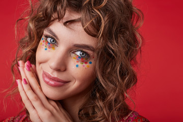 Positive pretty young female with romantic hairstyle and multicolored dots on her face looking at camera with tender smile and leaning chin on folded hands, standing over red background