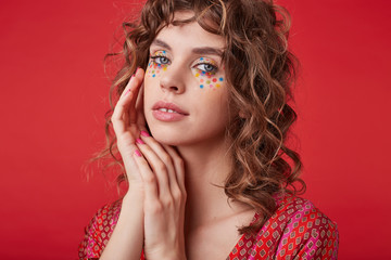 Delicate pretty lady with romantic hairstyle and multicolored dots on her face wearing motley patterned top while posing over red background, folding hands near face and looking gently to camera
