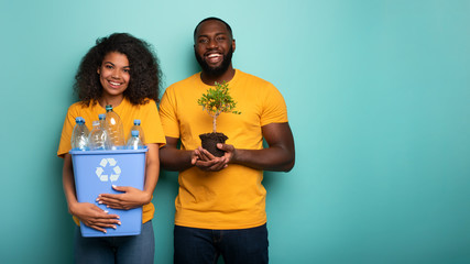 Happy couple hold a plastic container and a small tree over a light blue color. Concept of...