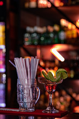 bebida alcohólica roja en una copa de martini con pedazos de limón y al lado un .vaso con pitillos en un bar 