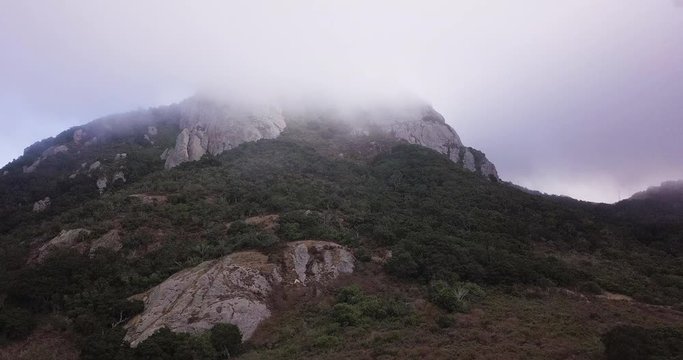 Ascending aerial shot of a green, rocky mouintain at Morro Bay in California, USA with a misty, foggy peak - Left Truck