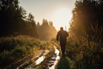 Vintage hunter idzie leśną drogą. Łowca karabinów w pięknym zachodzie lub wschodzie słońca. Karabin myśliwy w lesie - 297435129