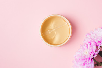 Golden patches on a pink pastel background next to fresh chrysanthemum flowers. Natural flower extract cosmetics, beauty and fashion. Patches for moisturizing in a jar, flat lay, top view