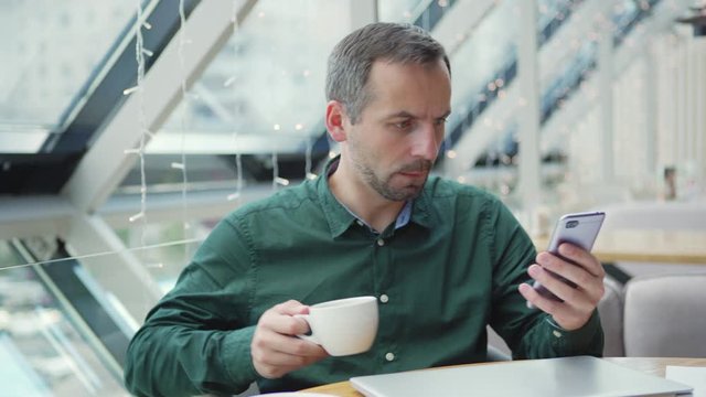 Serious businessman sipping hot drink and reading message on smartphone then starting to make corrections in data on laptop and exhaling in relief while working on project in cafe