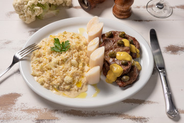 Risotto with filet mignon. Risoto and beef with capers and mustard in a white plate on wooden white background. Soft light. Italian food.